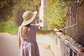 Little girl in straw hat reaching for flowers on wooden fence