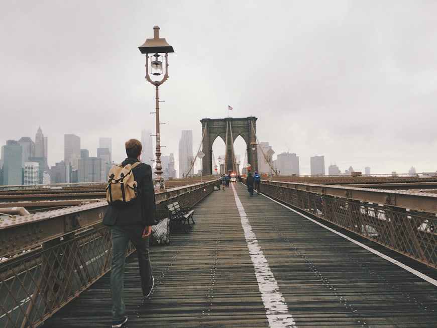Man with backpack walking on Brooklyn Bridge towards Manhattan skyline