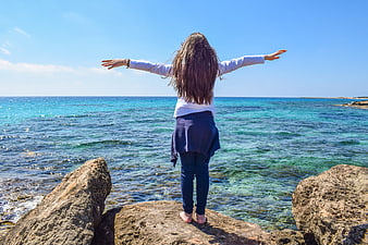Woman with arms outstretched standing on rocks by turquoise sea
