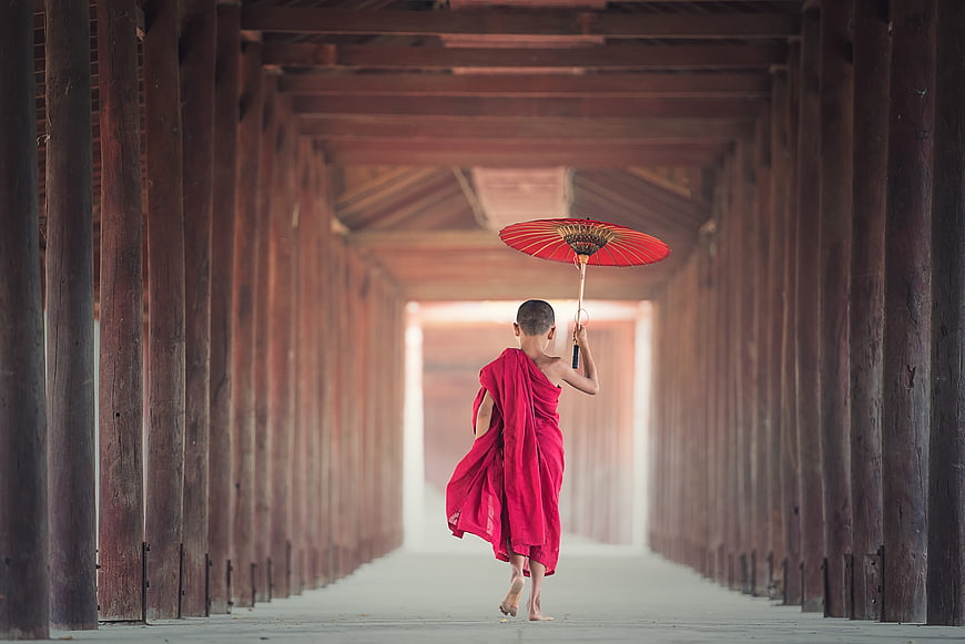Young Buddhist Monk Walking with Red Umbrella in Wooden Corridor