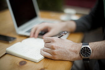 Man writing in notebook with laptop and watch on wooden table