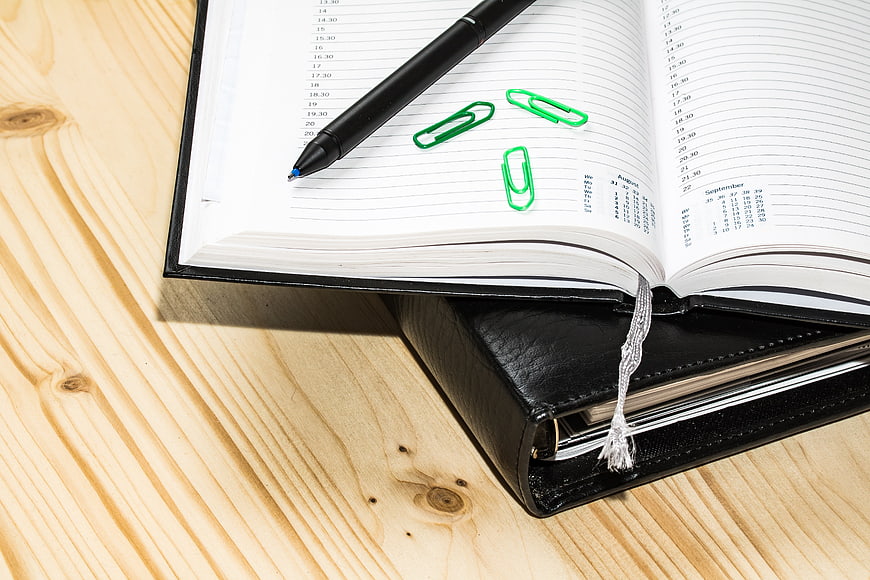 Open planner with black pen and green paperclips on wooden desk