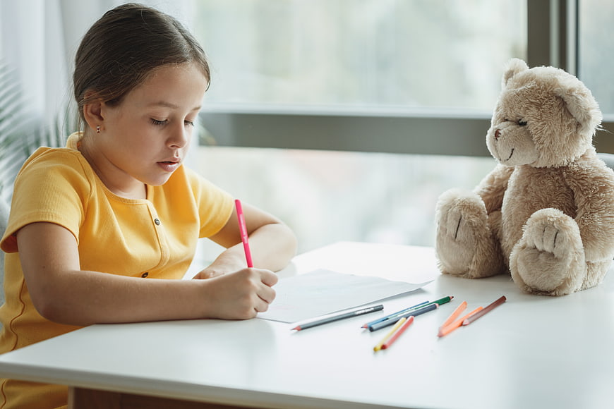 Young girl drawing at desk with teddy bear