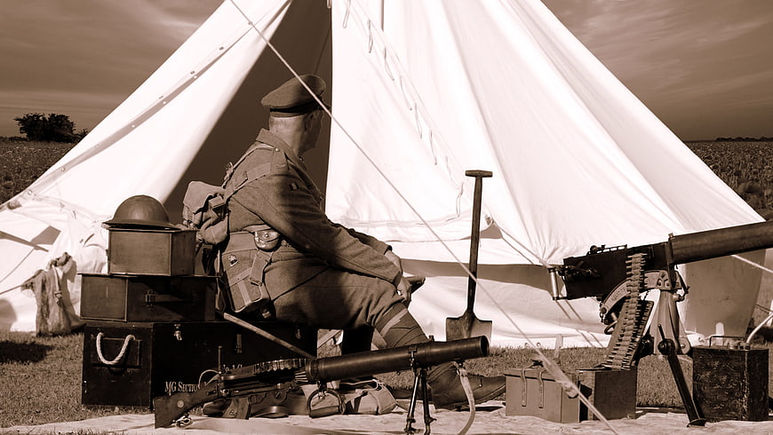 Sepia-toned WWI reenactor sitting by military tent with machine gun