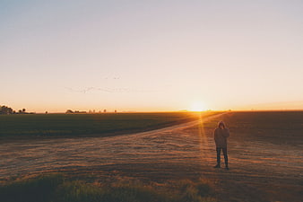 Lone figure standing on dirt road at sunset in open field