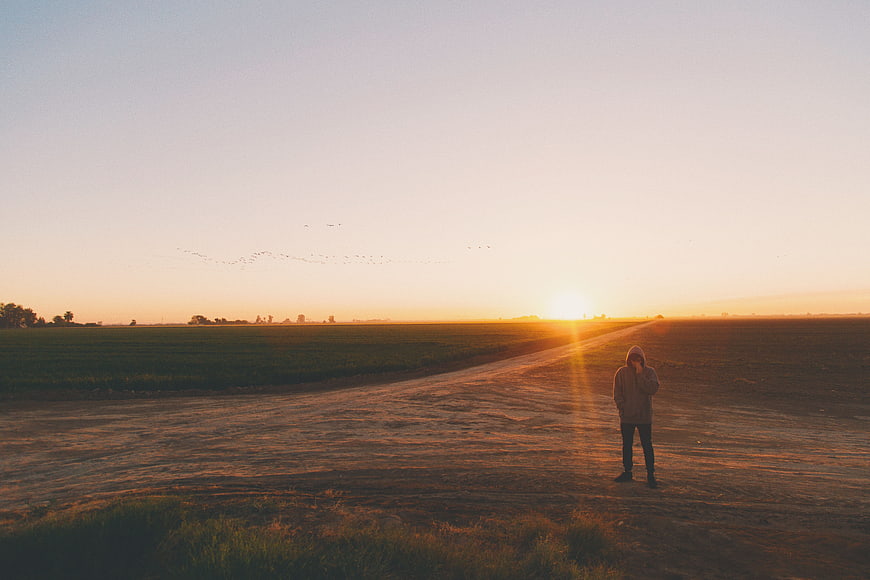 Lone figure standing on dirt road at sunset in open field