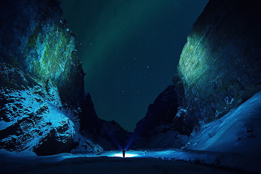 Silhouette of Person with Flashlight in Snowy Canyon Under Aurora Borealis