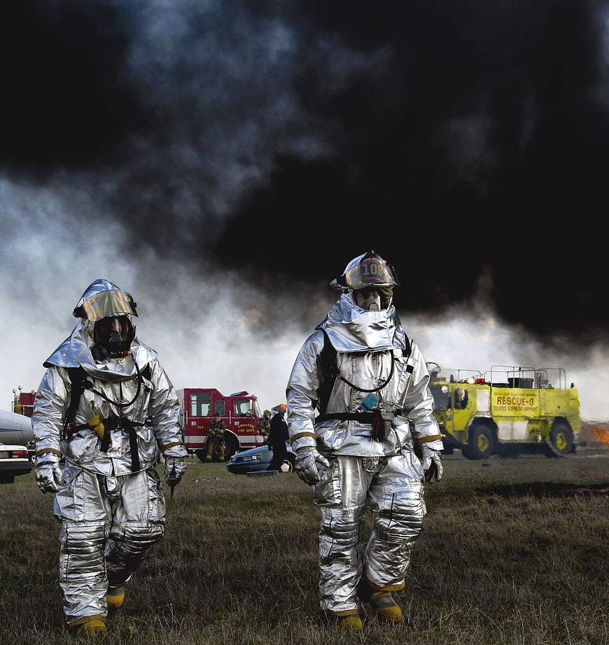 Firefighters in silver protective suits walking through smoke at a fire scene