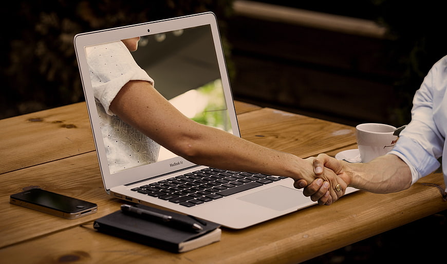 Handshake emerging from laptop screen on wooden table