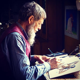 Senior male artist with long beard drawing in a dimly lit studio