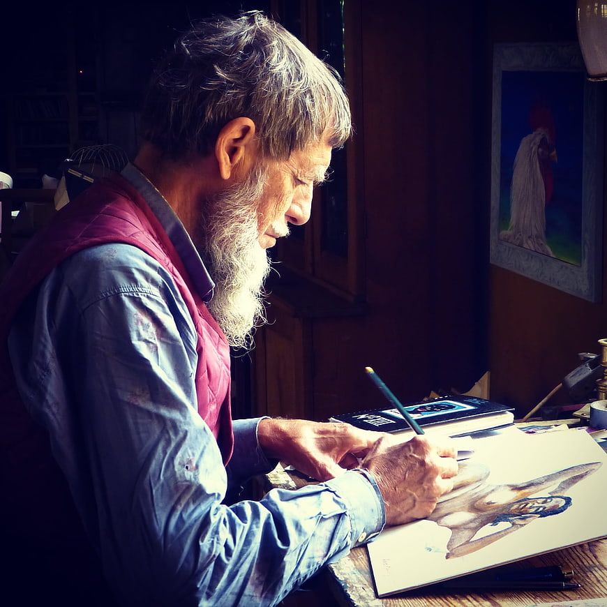Senior male artist with long beard drawing in a dimly lit studio