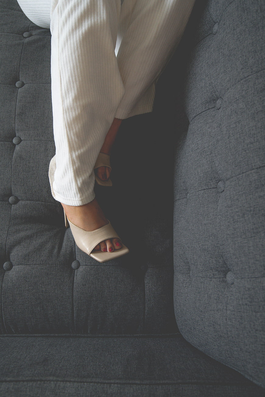 Woman's legs in white trousers and beige heels on gray sofa