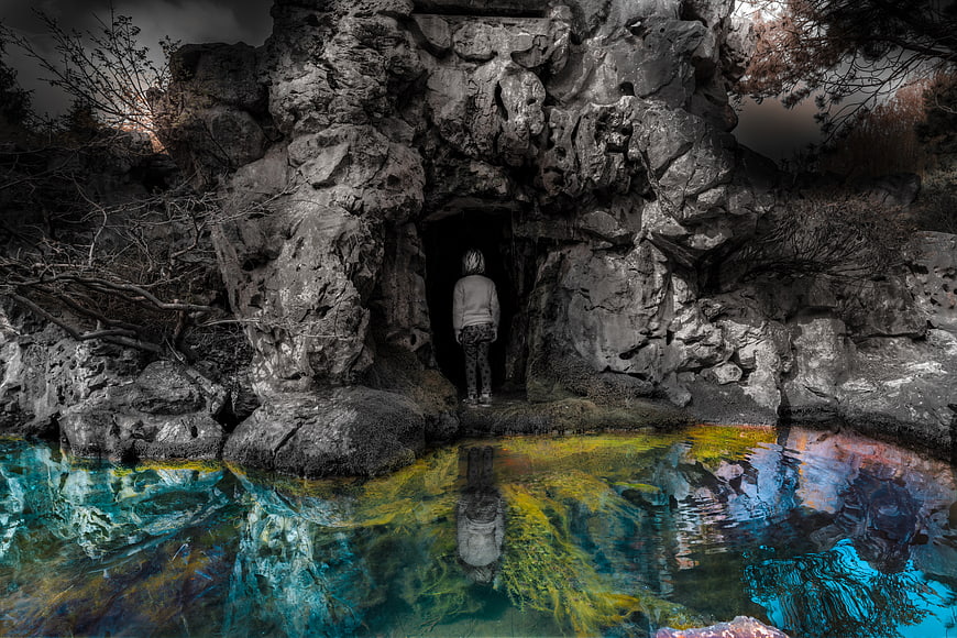 Mysterious figure standing in cave entrance with colorful water reflection