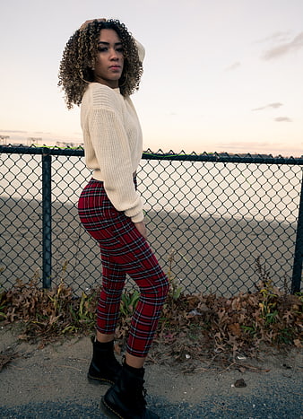 Stylish woman in plaid pants posing by chain-link fence