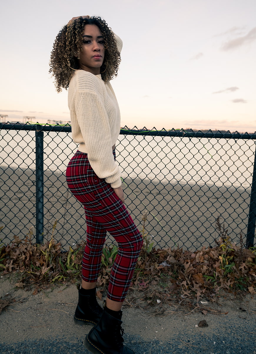 Stylish woman in plaid pants posing by chain-link fence