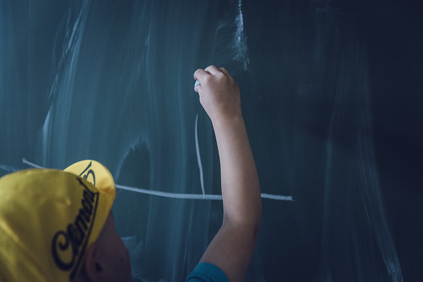 Child Writing on Chalkboard with Yellow Cap