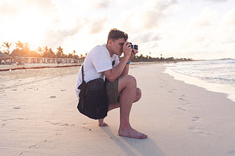 Young male photographer crouching on beach taking photo at sunset