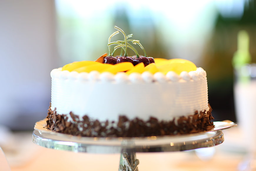 White birthday cake with yellow fruit and cherries on silver stand