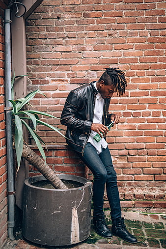 Stylish man with dreadlocks sitting against brick wall
