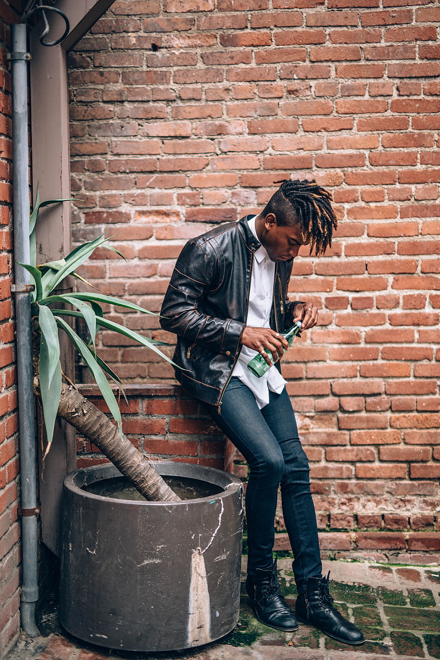 Stylish man with dreadlocks sitting against brick wall