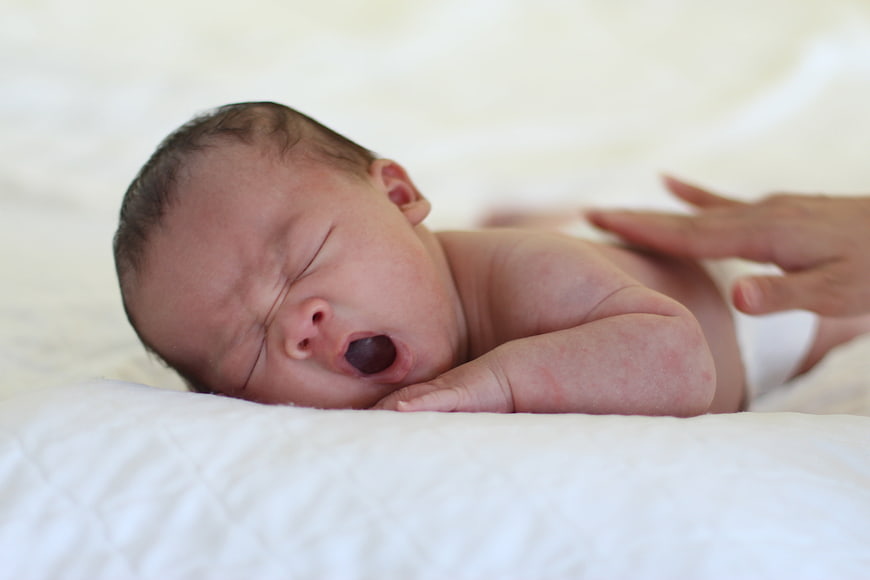 Newborn Baby Yawning On White Bedding With Parent Hand