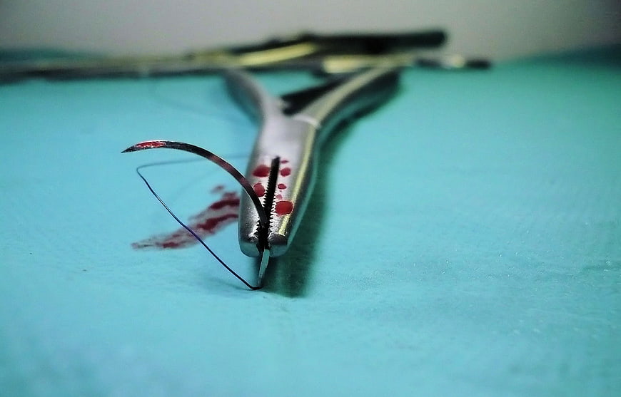 Blood-stained surgical needle holder and curved needle on teal surface