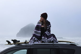 Woman wrapped in blanket sitting on car roof with surfboard by foggy ocean