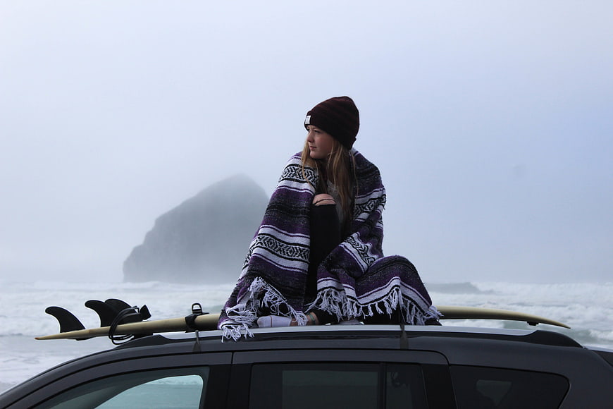Woman wrapped in blanket sitting on car roof with surfboard by foggy ocean