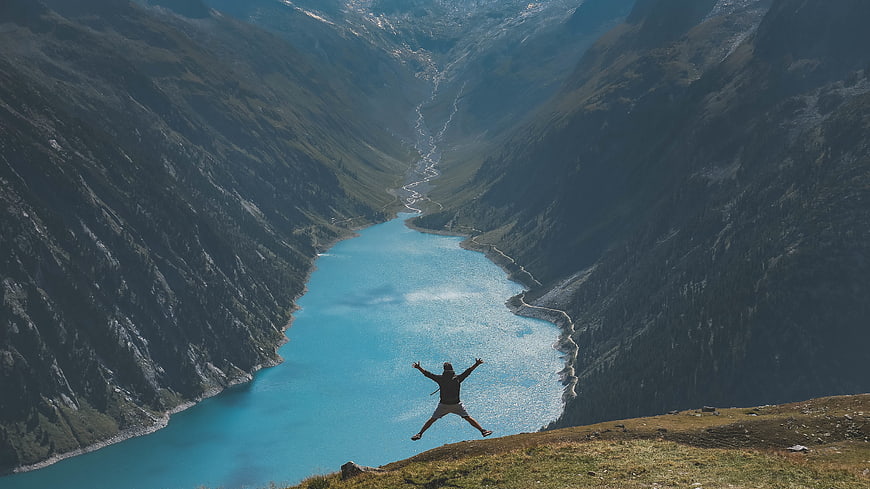 Man jumping with arms raised on mountain ridge overlooking turquoise lake