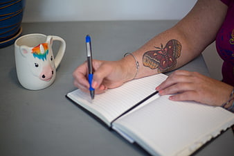 Woman writing in notebook with unicorn mug and tattoo