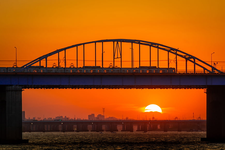 Silhouette of a steel arch bridge with a train against a vibrant orange sunset over the sea