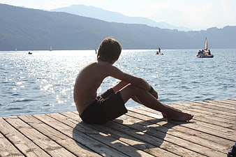 Boy sitting on wooden pier looking at lake with mountains