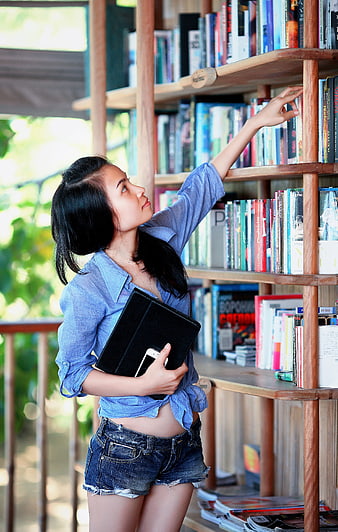 Young woman reaching for book on wooden shelf in library