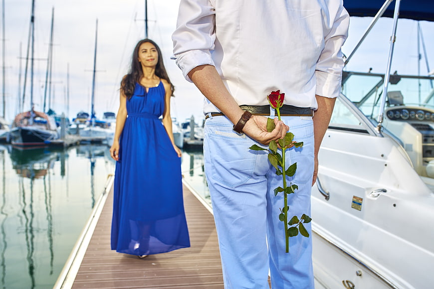 Man holding rose behind back at marina with woman