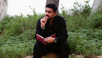 Indian man in black shirt sitting outdoors holding red book