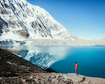 Man standing by turquoise glacial lake with snow-capped mountain reflection