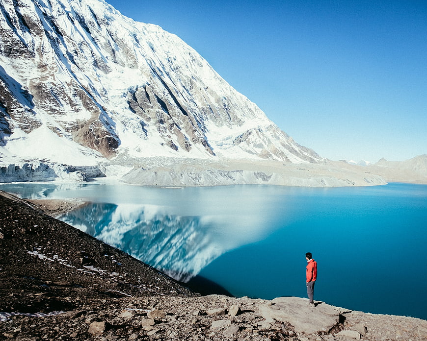 Man standing by turquoise glacial lake with snow-capped mountain reflection