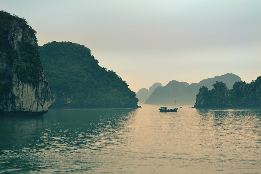 Lone boat sailing through misty limestone karst islands at dawn