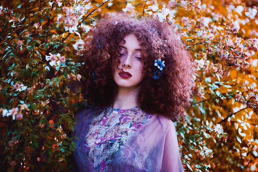 Woman with curly red hair in floral dress amidst blooming bushes