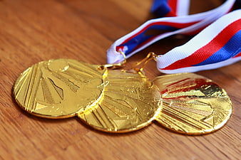 Three Gold Sports Medals With Red White And Blue Ribbons On Wooden Table