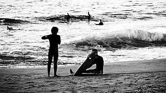 Black and white silhouette of surfers on a sandy beach with ocean waves