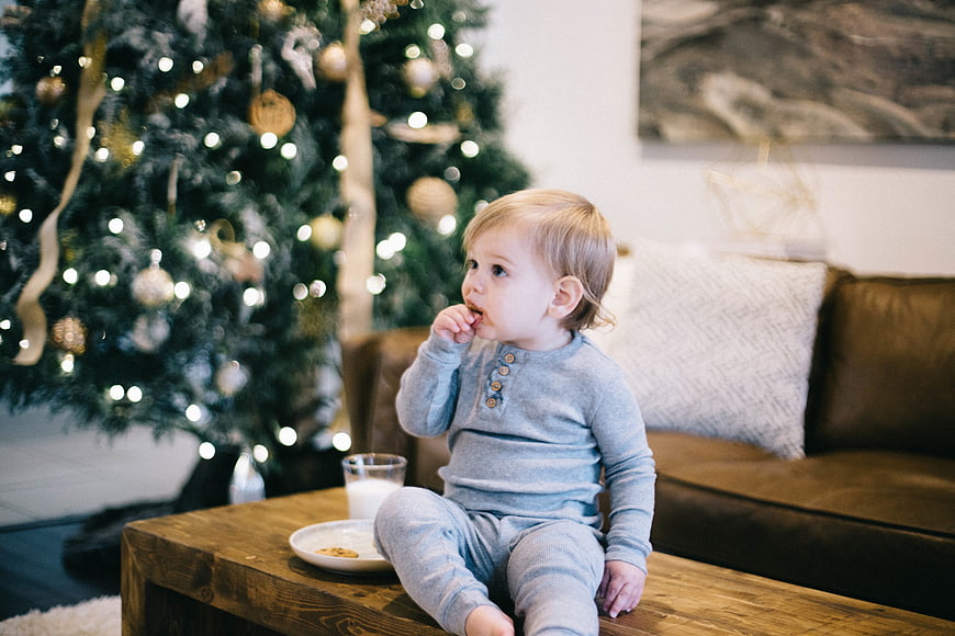 Toddler boy eating cookies and drinking milk near Christmas tree