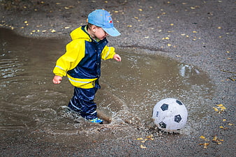 Young boy in rain gear playing with soccer ball in puddle