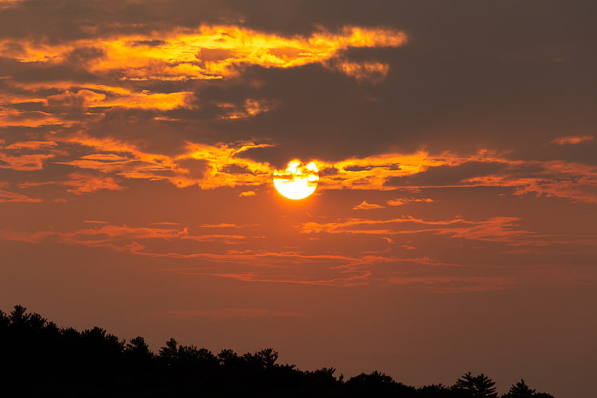 Vibrant orange sunset with sun behind clouds over tree silhouette