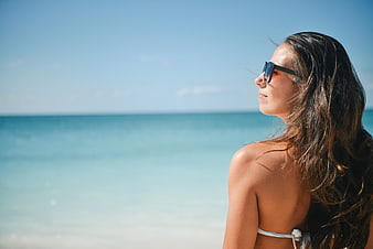 Young woman in bikini and sunglasses looking at the ocean on a sunny beach