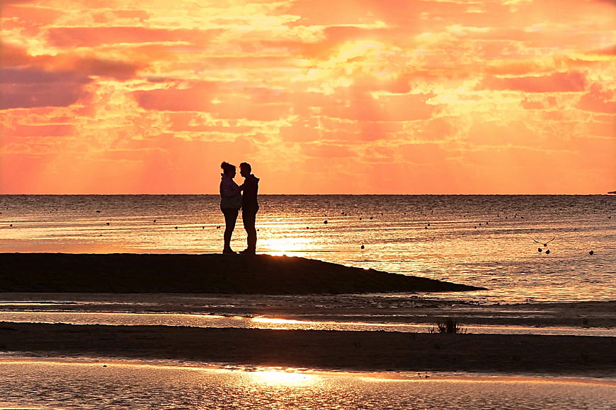 Silhouetted couple standing on a sandbar during a vibrant orange sunset