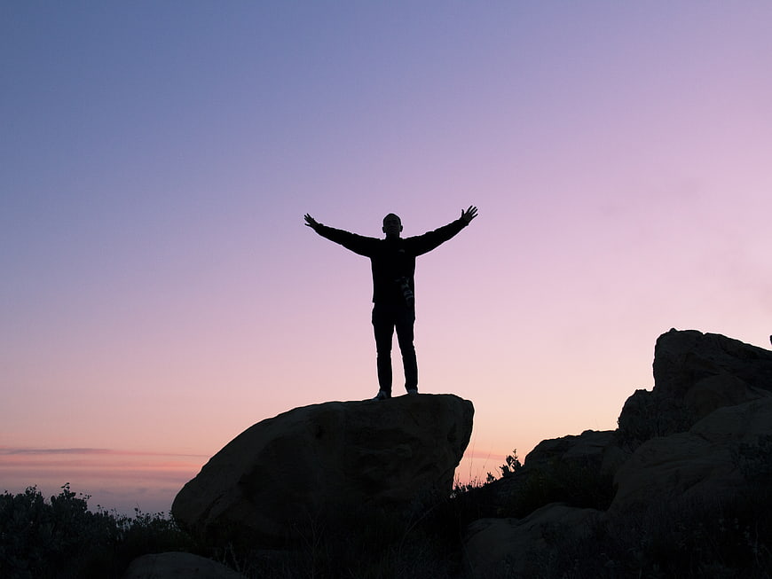 Silhouette of Man Standing on Rock with Arms Raised at Sunset