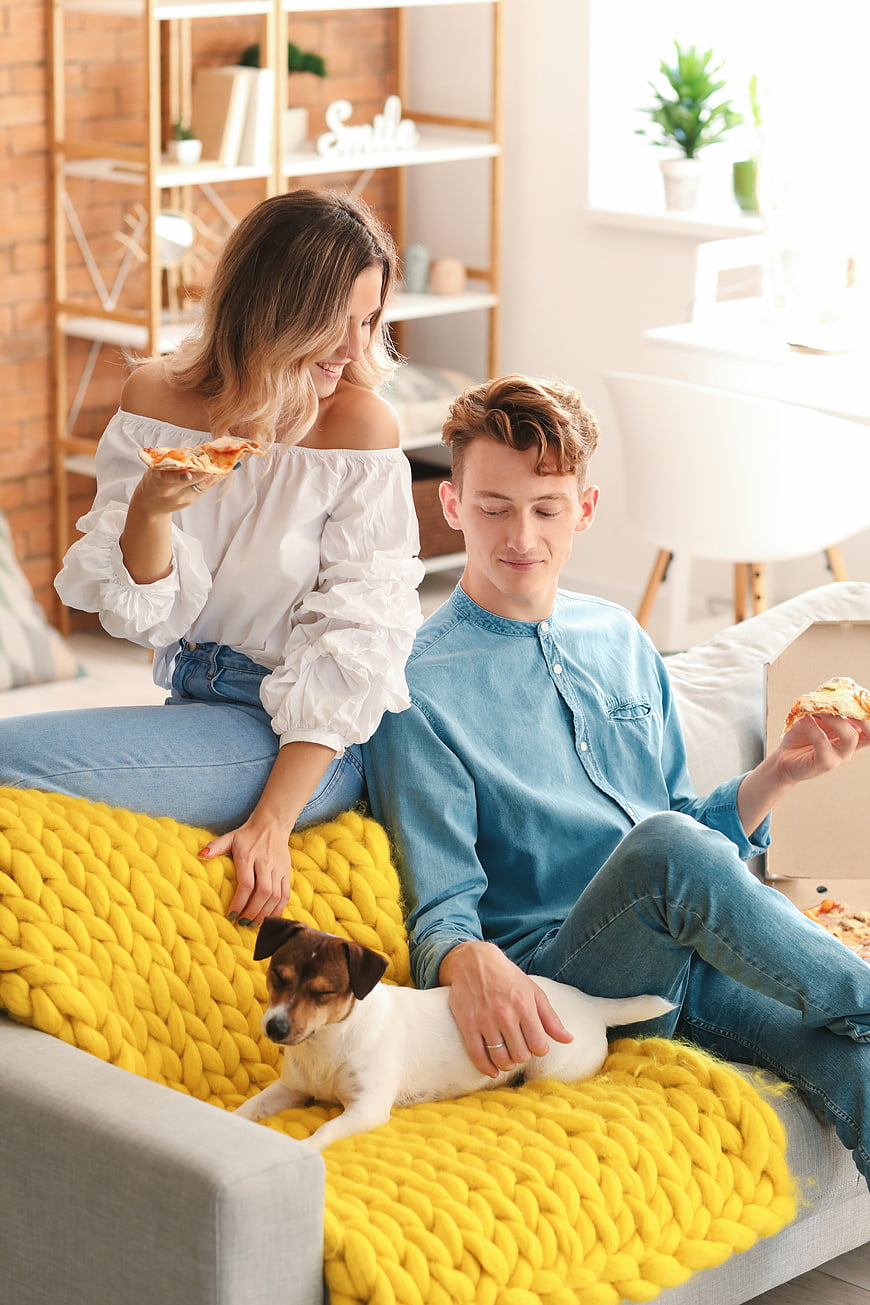 Happy young couple eating pizza on sofa with dog