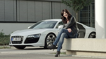 Smiling woman sitting on ledge next to white Audi R8 sports car
