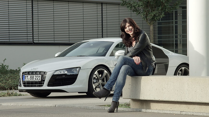 Smiling woman sitting on ledge next to white Audi R8 sports car